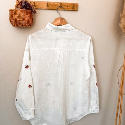 Camisa branca fina com bordados vermelhos nas mangas num cabide de madeira e cesta de verga com flores secas