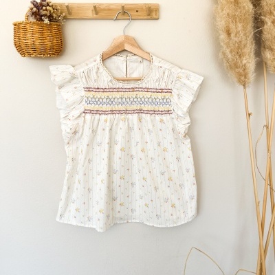 Blusa branca florida com detalhes coloridos pendurada em cabide de madeira numa parede clara com decoração seca