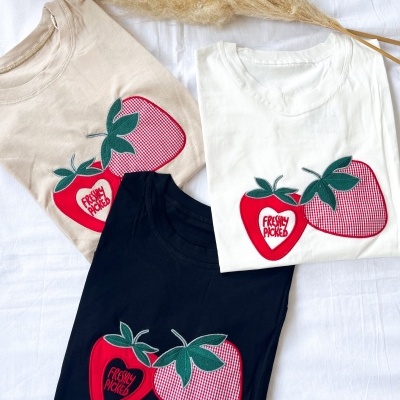 Três t-shirts dobradas com estampas de morangos vermelhos e texto FRESHLY PICKED
