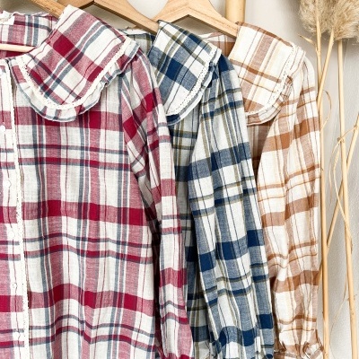 Três camisas xadrez coloridas com golas largas em cabides de madeira num suporte de bambu