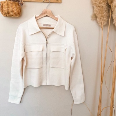 Casaco branco com fecho de correr e bolsos pendurado numa parede com decoração seca
