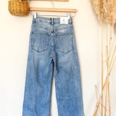 Calças jeans azuis claras wide leg penduradas numa cabide de madeira com parede branca e decoração natural