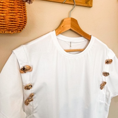 T-shirt branca com botões dourados decorativos pendurada num cabide de madeira