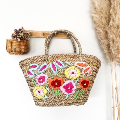 Mala de palha com flores bordadas pendurada numa parede branca ao lado de flores secas.