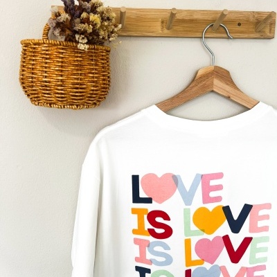 T-shirt branca com estampa colorida LOVE IS LOVE, pendurada em cabide de madeira na parede
