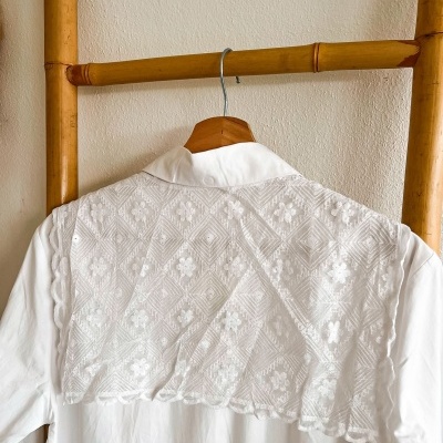 Camisa branca com bordado floral pendurada em cabide de madeira numa estrutura de madeira.