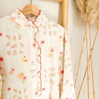 Blusa floral clara com folhos pendurada em cabide de madeira num suporte de bambu