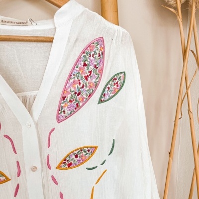 Blusa branca com aplicação de patches florais coloridos e bordados, pendurada em cabide de madeira numa parede clara com plantas secas