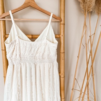 Vestido branco texturizado pendurado em cabide de madeira com estrutura de bambu e plantas secas ao lado