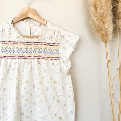 Blusa feminina branca com flores pequenas e listras azuis pendurada em cabide de madeira.