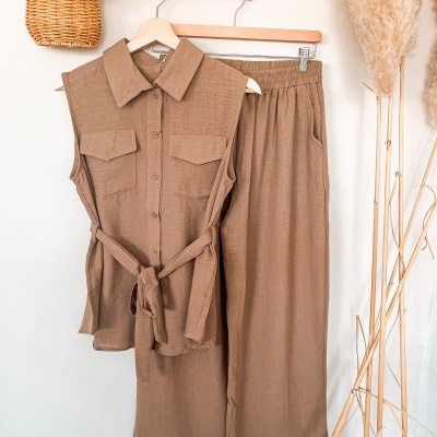 Conjunto de camisa sem mangas e calças castanhas pendurado na parede junto a cestinho de vime com flores e plantas secas.