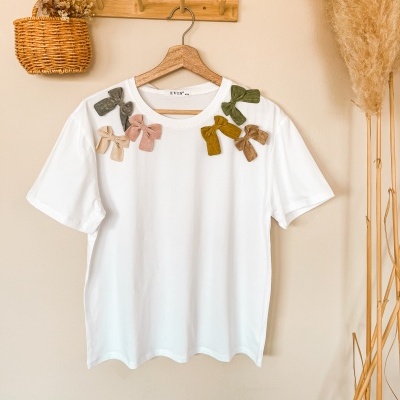 T-shirt branca com laços coloridos pendurada em cabide de madeira numa parede bege