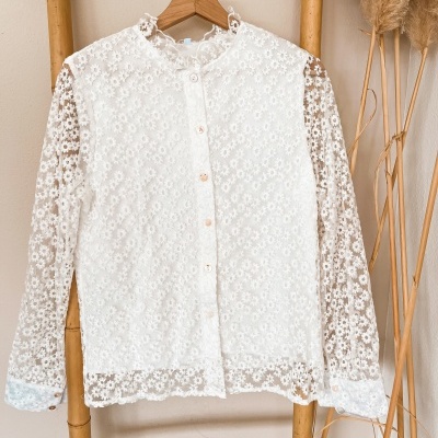 Camisa branca de renda floral pendurada em cabide numa estrutura de bambu com plantas secas ao lado