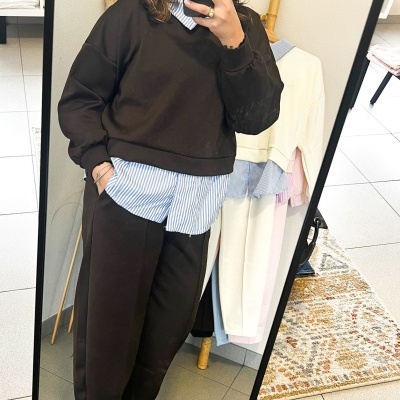 Pessoa vestindo conjunto castanho escuro com camisa azul às riscas e ténis brancos, a olhar ao espelho numa divisão com cabide de roupa.