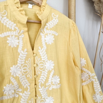 Blusa amarela com bordados florais brancos pendurada em cabide de madeira