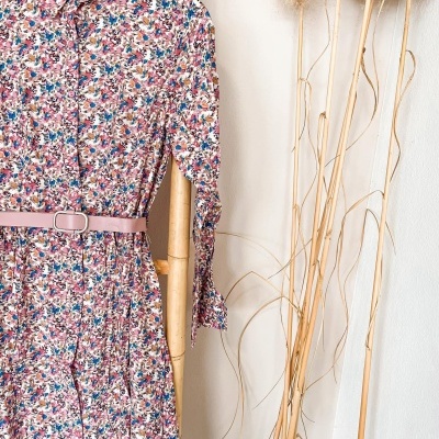 Vestido floral com cinto rosa pendurado em estrutura de madeira ao lado de ramos secos.