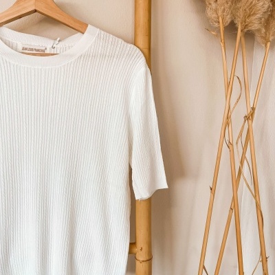 Camisola branca de malha canelada pendurada em cabide numa estrutura de bambu com planta decorativa ao lado