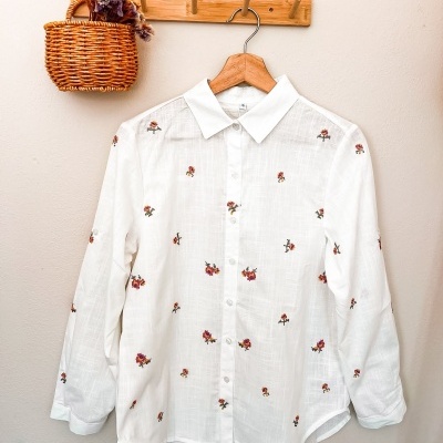 Camisa branca bordada pendurada num cabide de madeira com cesta de vime à esquerda