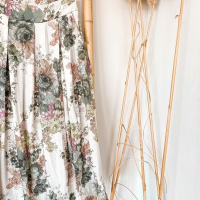 Saia floral colorida pendurada em cabide de madeira junto a hastes secas contra parede branca.