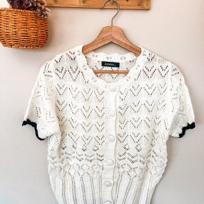 Camisola branca de malha rendada com botões em cabide de madeira e cesto com flores secas.