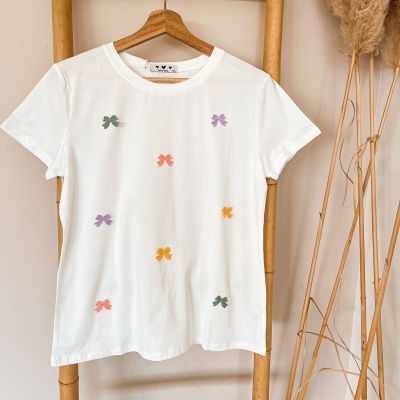 T-shirt branca com laços bordados coloridos pendurada em cabide de madeira