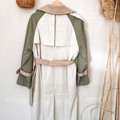 Casaco trench coat comprido em bege, verde e camel pendurado em gancho de madeira com cesto de vime e plantas secas ao lado