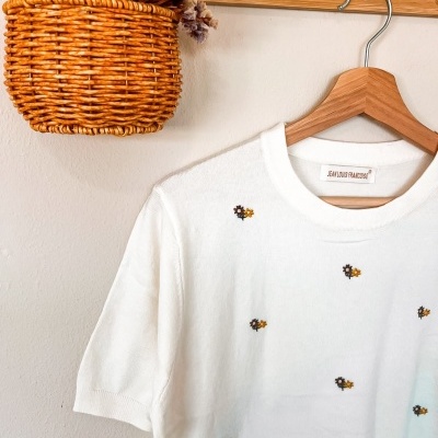 camisola branca com flores bordadas pendurada em cabide de madeira numa parede branca com cesta de verga com flores secas