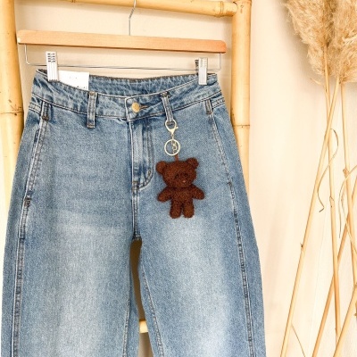 Calças de ganga azuis claras com chaveiro artesanal em forma de ursinho castanho penduradas num cabide de madeira.