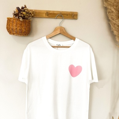 T-shirt branca com coração rosa pendurada num cabide de madeira numa parede creme com decoração natural