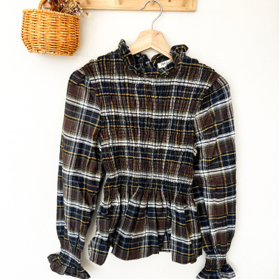Blusa xadrez castanha com mangas compridas pendurada em cabide de madeira e cesto de verga com flores secas