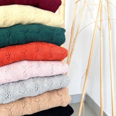 Pilhas de camisolas de malha coloridas com padrão e textura visível, empilhadas numa cadeira branca ao lado de estrutura de bambu.