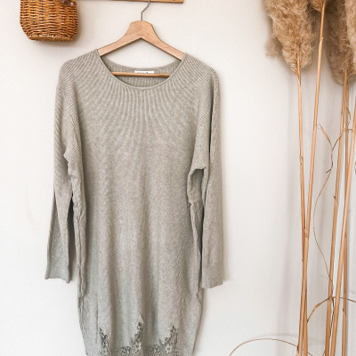 Vestido cinzento de malha com renda pendurado numa parede branca com decoração de flores secas e cesto de vime
