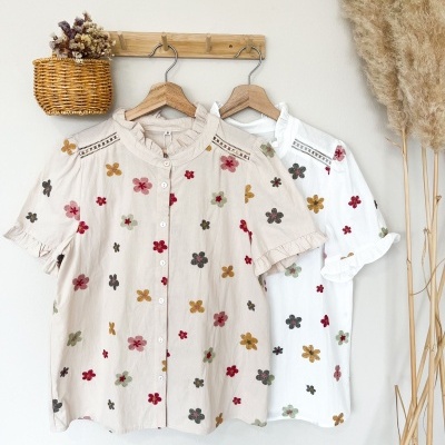 Duas blusas florais em cabides de madeira num suporte de parede junto a uma cesta de verga com flores secas.