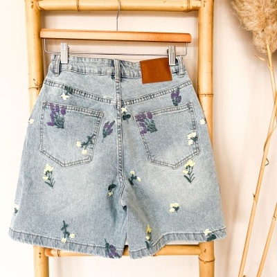 Calções de ganga azul clara com flores bordadas e etiqueta de couro, pendurados num cabide de madeira numa estrutura de bambu.