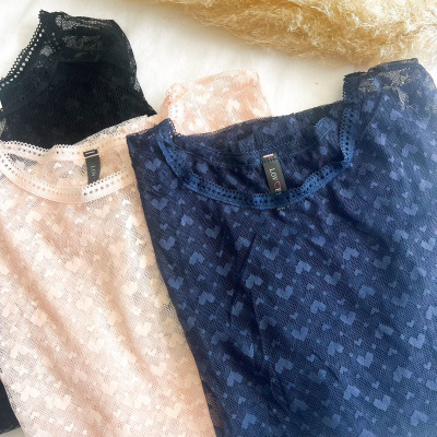 Três blusas de renda com padrão de corações nas cores preta, rosa claro e azul escuro sobre superfície branca e palha seca ao fundo
