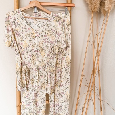 Conjunto feminino floral pendurado em cabides de madeira numa estrutura de bambu, ao lado de ramos secos decorativos.