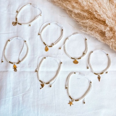 Pulseiras brancas com contas douradas e pendentes em metal dourado sobre tecido branco com palha decorativa