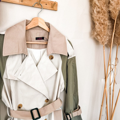 Casaco trench coat bicolor creme e verde pendurado numa parede branca ao lado de ramos secos.