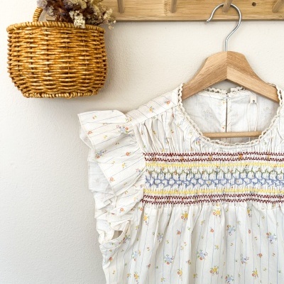 vestido branco floral com bordados coloridos num cabide de madeira junto a um cesto de vime com flores secas