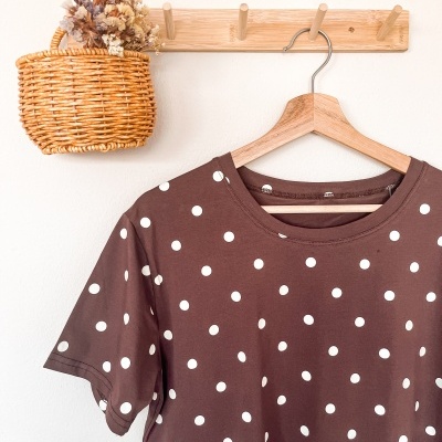 T-shirt castanha de bolinhas brancas pendurada em cabide de madeira com cesto de verga e flores secas junto ao suporte de madeira.