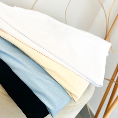 Quatro t-shirts dobradas em branco, creme, azul claro e preto em superfície clara