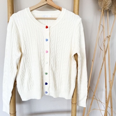 Casaco de malha branco com botões coloridos em forma de coração pendurado numa estrutura de bambu