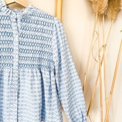 Blusa feminina azul claro com padrão xadrez e corações, botões brancos e detalhes franzidos pendurada num cabide de madeira.