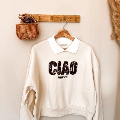 Sweatshirt creme com gola branca e impressão CIAO Amore pendurada num cabide de madeira em gancho de madeira numa parede creme com cesto de vime com flores secas ao lado.