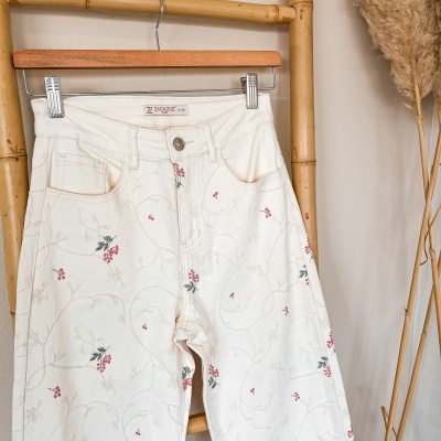 Calças de ganga brancas bordadas com flores penduradas em cabide num suporte de bambu