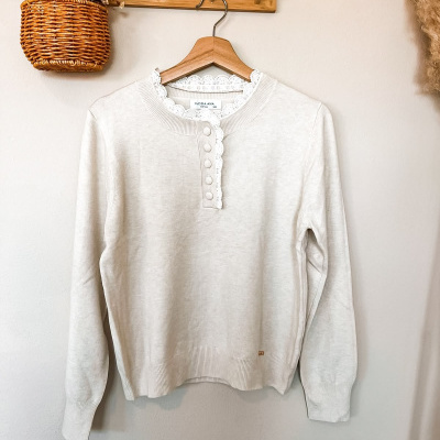 Camisola creme com renda branca na gola e botões forrados, pendurada num cabide de madeira na parede com cesto de vime e flores secas
