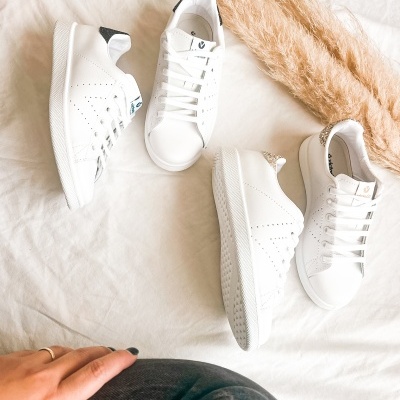 Quatro pares de sapatilhas brancas sobre tecido branco com folhas secas decorativas e mão numa perna com calças escuras