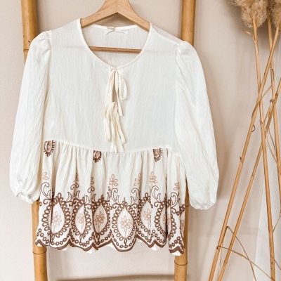 Blusa branca com bordados castanhos pendurada num cabide de madeira