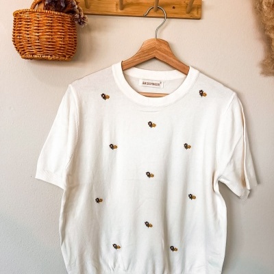 Camisola branca com flores bordadas pendurada num cabide de madeira com cesto de verga com flores secas ao lado.