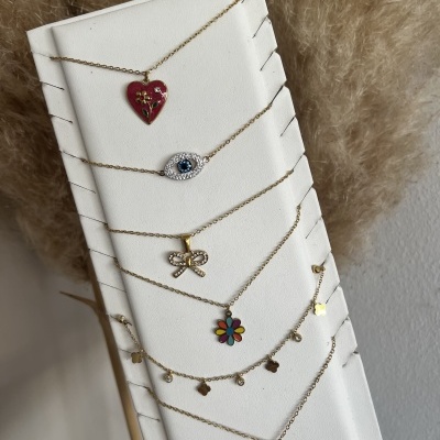 Colares dourados com pendentes coloridos em suporte branco e planta seca ao fundo.